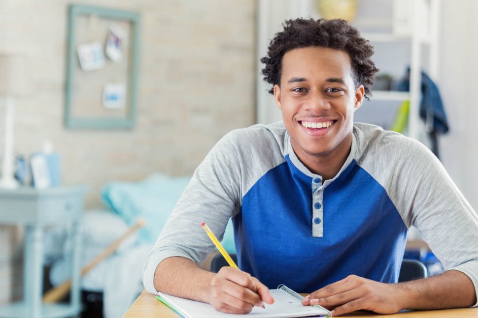Cheerful African American teenage boy studies in his room. He is smiling at the camera. He is writing in a spiral notebook.