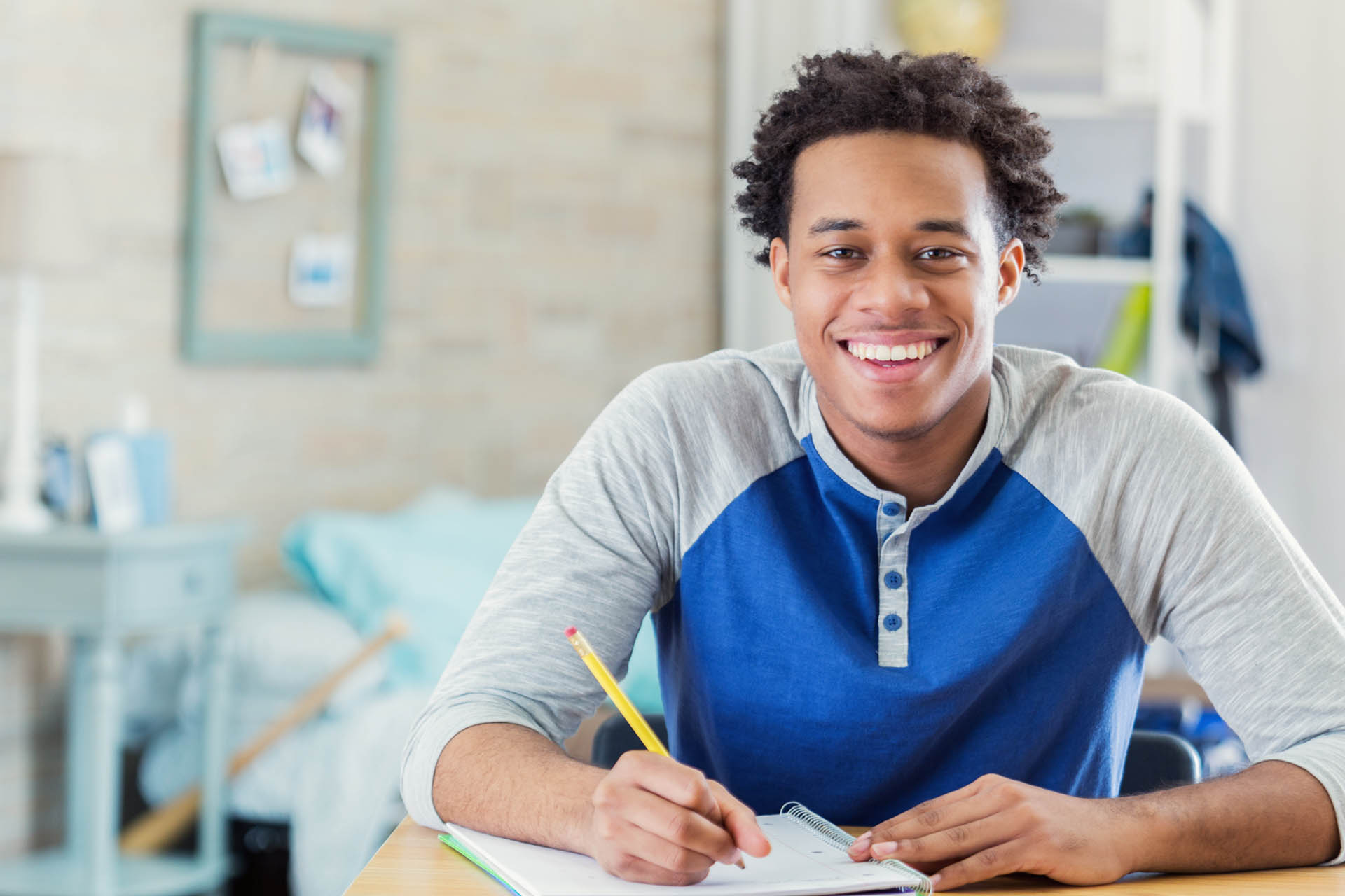 Cheerful African American teenage boy studies in his room. He is smiling at the camera. He is writing in a spiral notebook.